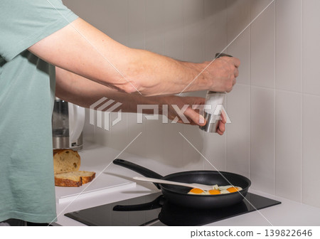 Mature man adds seasoning into frying pan during cooking process. Adult male prepares breakfast meal at kitchen stove in modern home interior. Everyday domestic food preparation routine.. 139822646