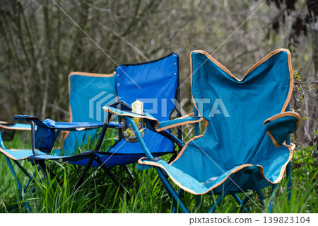 A row of blue foldable camping chairs with cup holders set up on a lush green lawn. Evoking feelings of relaxation, picnic, or outdoor event waiting to happen. A row of blue foldable camping chairs with cup holders set up on a lush green lawn. Evoking feelings of relaxation, picnic, or outdoor event waiting to happen. 139823104
