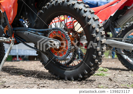 Close-up of a rear knobby tire, orange rear sprocket, and drive chain on an off-road motocross motorcycle outdoors. Detail view of a dirt bike rear wheel mechanism, drive chain, and knobby tire Close-up of a rear knobby tire, orange rear sprocket, and drive chain on an off-road motocross motorcycle outdoors. Detail view of a dirt bike rear wheel mechanism, drive chain, and knobby tire 139823117
