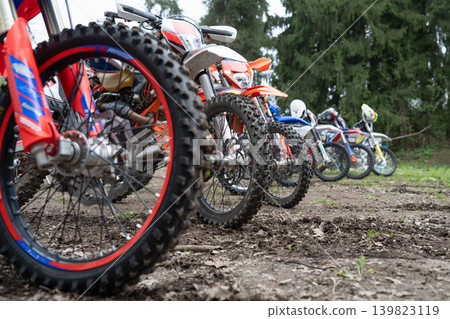 Close-up of motocross bike wheels with knobby tires lined up on a dirt track. Row of professional enduro motorcycles parked on muddy ground before the race. Close-up of motocross bike wheels with knobby tires lined up on a dirt track. Row of professional enduro motorcycles parked on muddy ground before the race. 139823119