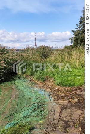 Polluted riverbank featuring stagnant green water, debris, and neglected natural surroundings. Documentary-style atmosphere highlights environmental issues. 139823357