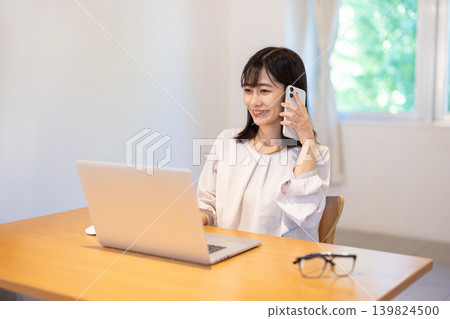 A young Japanese woman working on her computer while talking on her smartphone. A young Japanese woman working on her computer while talking on her smartphone. 139824500