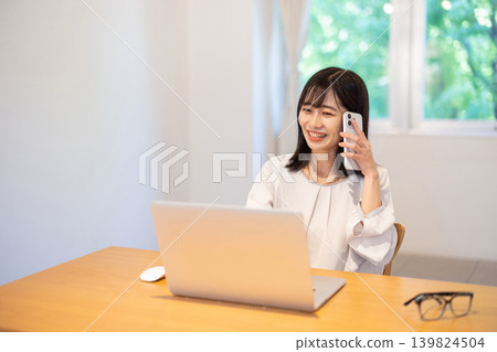 A young Japanese woman working on her computer while talking on her smartphone. A young Japanese woman working on her computer while talking on her smartphone. 139824504