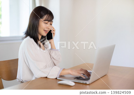 A young Japanese woman working on her computer while talking on her smartphone. A young Japanese woman working on her computer while talking on her smartphone. 139824509