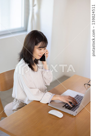 A young Japanese woman working on her computer while talking on her smartphone. 139824511