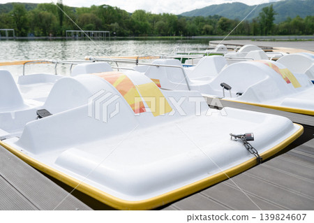 A row of modern white paddle boats parked at a wooden pier on a calm lake with green forest and mountains in the background. A row of modern white paddle boats parked at a wooden pier on a calm lake with green forest and mountains in the background. 139824607