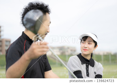 一位微笑的女士正在學習高爾夫球;這是她接受高爾夫教練揮桿指導的圖片。 一位微笑的女士正在學習高爾夫球;這是她接受高爾夫教練揮桿指導的圖片。 139824723