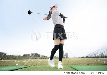 Image of a young, smiling woman swinging a driver at a golf driving range. Nice shot! Image of a young, smiling woman swinging a driver at a golf driving range. Nice shot! 139824997
