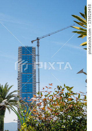 A modern high-rise building under construction with a tall crane, against a clear sky A modern high-rise building under construction with a tall crane, against a clear sky 139828458