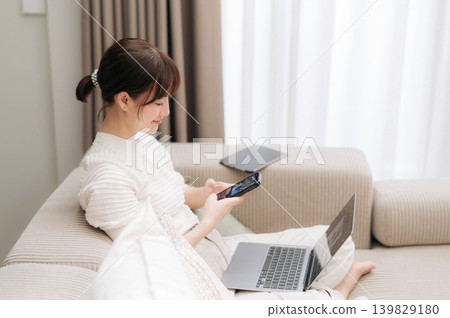 Happy beautiful young asian woman sitting comfortably on beige sofa using modern smartphone device and laptop computer at home feeling relaxed while browsing internet on lazy weekend morning Happy beautiful young asian woman sitting comfortably on beige sofa using modern smartphone device and laptop computer at home feeling relaxed while browsing internet on lazy weekend morning 139829180