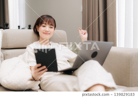 Happy young asian woman relaxing on comfortable sofa at home in living room while looking at tablet screen and using laptop computer enjoying cozy weekend leisure time Happy young asian woman relaxing on comfortable sofa at home in living room while looking at tablet screen and using laptop computer enjoying cozy weekend leisure time 139829183
