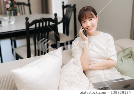 Happy young asian woman sitting on comfortable sofa talking on smartphone using laptop computer smiling bright modern living room home office communication freelance lifestyle telecommute Happy young asian woman sitting on comfortable sofa talking on smartphone using laptop computer smiling bright modern living room home office communication freelance lifestyle telecommute 139829184