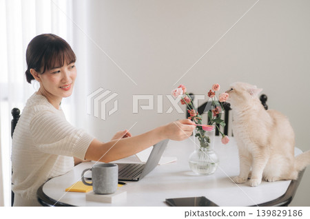 Joyful young asian woman smiling bright feeding fluffy white pet cat at home table with laptop computer feeling deep affection enjoying relaxed remote work lifestyle inside cozy room 139829186