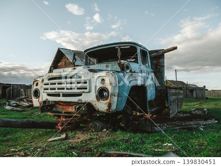 An old blue truck rests with wood logs under its front in the green field. Nearby, a wooden house stands in disrepair. An old blue truck rests with wood logs under its front in the green field. Nearby, a wooden house stands in disrepair. 139830451