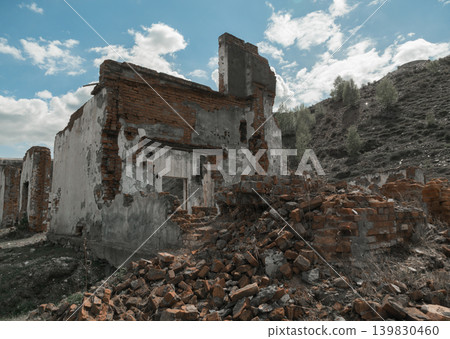 A broken-down building stands in a field surrounded by scattered bricks. The area shows signs of neglect and decay under a cloudy sky with visible hills. A broken-down building stands in a field surrounded by scattered bricks. The area shows signs of neglect and decay under a cloudy sky with visible hills. 139830460