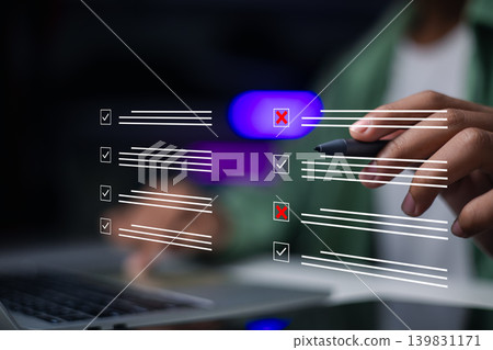 Hand using stylus pen to evaluate checklist on laptop. Businessman checking boxes with tick and cross marks for quality control, survey, and business audit assessment. 139831171