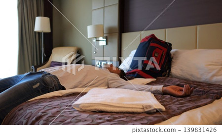 Exhausted man lying on bed in modern hotel room after long business trip. Tired young businessman relaxing in comfortable accommodation interior after work, travel and stressful daily routine Exhausted man lying on bed in modern hotel room after long business trip. Tired young businessman relaxing in comfortable accommodation interior after work, travel and stressful daily routine 139831426