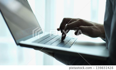Male hand of young businessman typing text on laptop keyboard near window in modern office. Professional manager working on computer with natural light, showing productivity and communication Male hand of young businessman typing text on laptop keyboard near window in modern office. Professional manager working on computer with natural light, showing productivity and communication 139831428
