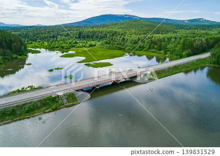 Concrete bridge extends over a wide river, connecting forested shores and leading to distant green mountains 139831529