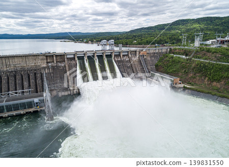 Aerial view of concrete dam releasing water into river on cloudy day. Water discharge at hydroelectric power plant. Aerial view of concrete dam releasing water into river on cloudy day. Water discharge at hydroelectric power plant. 139831550