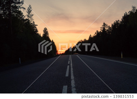 A long, empty asphalt road with white double lines extends through a dark forest at sunset A long, empty asphalt road with white double lines extends through a dark forest at sunset 139831553