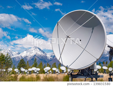 Many white satellite dishes, including a prominent one in front, are positioned in a grassy field Many white satellite dishes, including a prominent one in front, are positioned in a grassy field 139831580