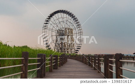 A large wooden water wheel stands tall beside a wooden boardwalk path winding through a green wetland. 139831617