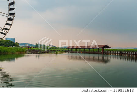 Wooden covered boardwalk stretching across serene lake with a large water wheel on a cloudy day Wooden covered boardwalk stretching across serene lake with a large water wheel on a cloudy day 139831618