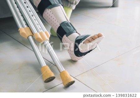 Lower leg of a person wearing a medical walking boot with exposed toes. Aluminum crutches with yellow tips stand next to it on a light tiled floor, indicating an injury or recovery process. Lower leg of a person wearing a medical walking boot with exposed toes. Aluminum crutches with yellow tips stand next to it on a light tiled floor, indicating an injury or recovery process. 139831622