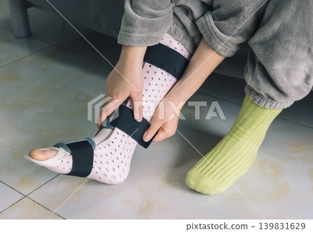 Close up of a person adjusting a medical ankle splint on their injured foot indoors. Close up of a person adjusting a medical ankle splint on their injured foot indoors. 139831629