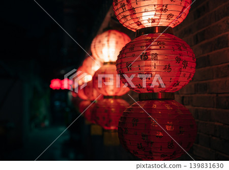 Bright red Chinese lanterns, decorated with gold characters, illuminate a dark brick wall at night 139831630