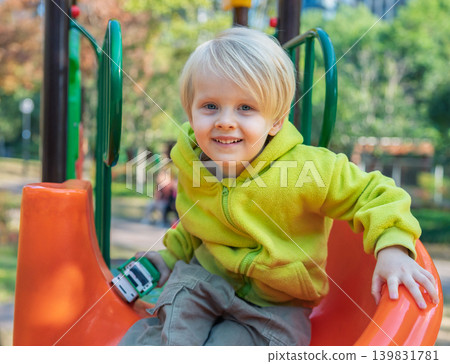 A happy blonde toddler boy sits on a bright orange slide, holding a small toy in his hand. He smiles widely in a park surrounded by colorful autumn trees A happy blonde toddler boy sits on a bright orange slide, holding a small toy in his hand. He smiles widely in a park surrounded by colorful autumn trees 139831781