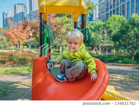 A happy blonde toddler boy sits on a bright orange slide, holding a small toy in his hand. He smiles widely in a park surrounded by colorful autumn trees A happy blonde toddler boy sits on a bright orange slide, holding a small toy in his hand. He smiles widely in a park surrounded by colorful autumn trees 139831782