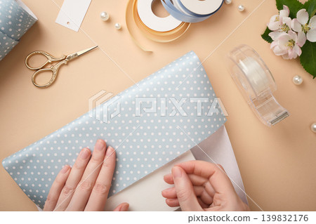 Woman's hands wrapping gift box with blue polka dot paper, gold scissors, tape dispenser, satin ribbons, pearl beads and flowers on beige background. Woman's hands wrapping gift box with blue polka dot paper, gold scissors, tape dispenser, satin ribbons, pearl beads and flowers on beige background. 139832176