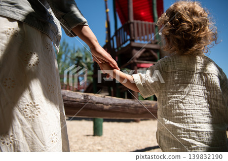 Mother and young son hold hands walking toward a wooden playground on a bright sunny day, conveying family bonding, childhood play, safety and outdoor summer togetherness. Mother and young son hold hands walking toward a wooden playground on a bright sunny day, conveying family bonding, childhood play, safety and outdoor summer togetherness. 139832190