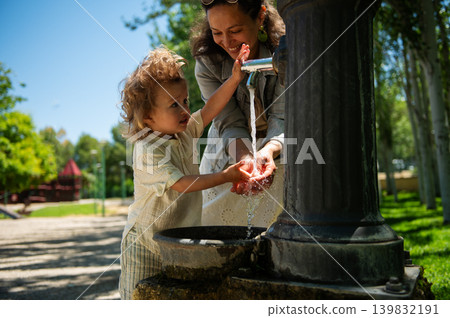 Mother helps her young son wash hands under a park fountain, sharing a joyful moment on a sunny day at the playground with family bonding and summer fun. Mother helps her young son wash hands under a park fountain, sharing a joyful moment on a sunny day at the playground with family bonding and summer fun. 139832191