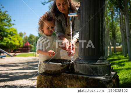 Mother and young son wash hands together at a park water fountain on a sunny day, sharing care, hygiene and playful outdoor moments in a children's playground. Mother and young son wash hands together at a park water fountain on a sunny day, sharing care, hygiene and playful outdoor moments in a children's playground. 139832192