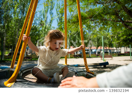 Toddler on a tire swing reaches for a parent's hand in a sunlit playground, capturing playful, curious and joyful family moments during a warm summer day outdoors. 139832194