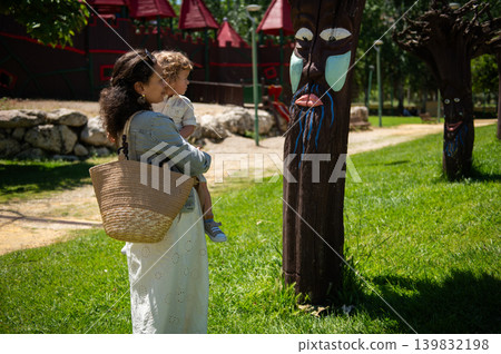 A mother holds her young son at a sunny kids park as they admire carved wooden totem faces, sharing a warm family moment of play, curiosity and parent-child bonding outdoors. A mother holds her young son at a sunny kids park as they admire carved wooden totem faces, sharing a warm family moment of play, curiosity and parent-child bonding outdoors. 139832198