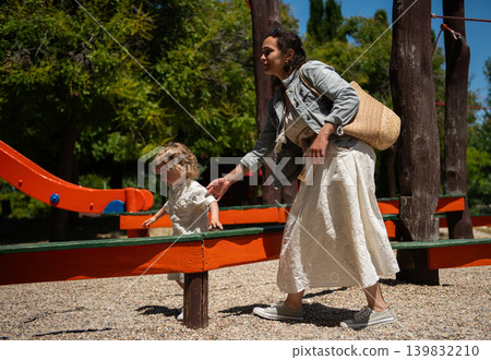 Mother and toddler son walk and play together at a sunny kids park, sharing a caring moment on playground equipment and gravel paths during a warm summer day. Mother and toddler son walk and play together at a sunny kids park, sharing a caring moment on playground equipment and gravel paths during a warm summer day. 139832210