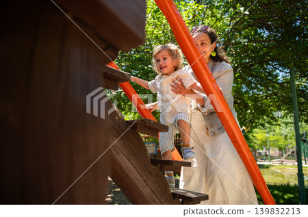 Mother and toddler son share joyful moment as she guides him up a wooden play structure in a leafy park on a bright sunny day, capturing warmth, trust and outdoor family fun. Mother and toddler son share joyful moment as she guides him up a wooden play structure in a leafy park on a bright sunny day, capturing warmth, trust and outdoor family fun. 139832213