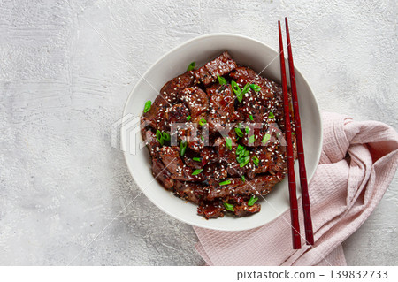 Crispy beef, with green onions and sesame seeds, in a bowl with chopsticks, on a textured surface, on a light background, no people Crispy beef, with green onions and sesame seeds, in a bowl with chopsticks, on a textured surface, on a light background, no people 139832733
