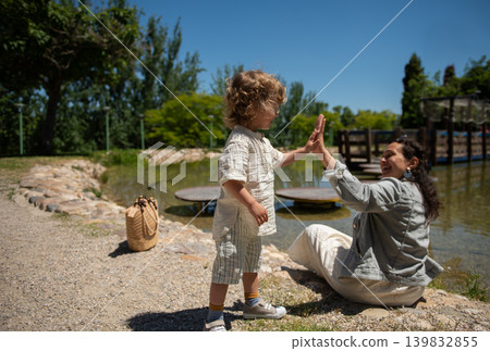 Mother and son high-five by a pond in a sunny kids park sharing a happy family moment Mother and son high-five by a pond in a sunny kids park sharing a happy family moment 139832855
