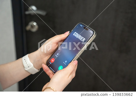 A woman receives a call from a courier standing at the front door of her apartment. Contactless home delivery service, online shopping logistics and modern urban lifestyle. A woman receives a call from a courier standing at the front door of her apartment. Contactless home delivery service, online shopping logistics and modern urban lifestyle. 139833662