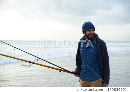 Relax, fishing and man walking on beach with rods, nature and outdoor holiday adventure. Ocean, tools and fisherman with pole, smile and cloudy sky with waves on winter morning vacation at sea. 139834361