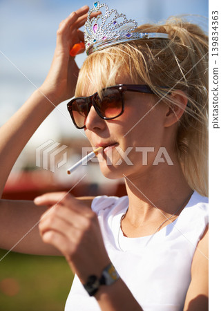 Crown, smoking and young woman with cigarette in nature at outdoor festival, event or party. Fun, tiara and female person with tobacco, sunglasses and casual fashion in outside park or field. 139834363