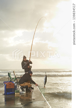 Fishing, men and net with fish at pier with rod, waves and relax on vacation, holiday and adventure. Friendship, people and bonding in morning with overcast, sky and nature for activity and hobby Fishing, men and net with fish at pier with rod, waves and relax on vacation, holiday and adventure. Friendship, people and bonding in morning with overcast, sky and nature for activity and hobby 139834517