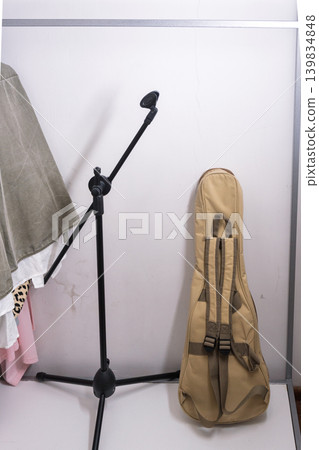 Vertical photo of a tan guitar gig bag and black microphone stand leaning against a white wall. Music equipment storage in a home wardrobe with hanging clothes. Vertical photo of a tan guitar gig bag and black microphone stand leaning against a white wall. Music equipment storage in a home wardrobe with hanging clothes. 139834848