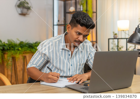 Indian man on remote learning uses laptop and notebook writes notes completes test quiz exam online Indian man on remote learning uses laptop and notebook writes notes completes test quiz exam online 139834870