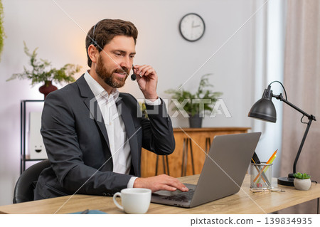 Middle-aged businessman service agent on headset listens and solves client issue while typing notes Middle-aged businessman service agent on headset listens and solves client issue while typing notes 139834935